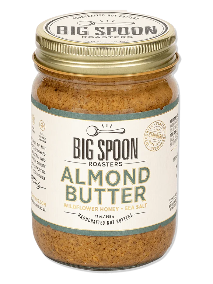 A glass jar of BIG SPOON ROASTERS - ALMOND BUTTER WILDFLOWER HONEY NUT BUTTER with a gold lid and a white label featuring branding and product details in black and gold text.