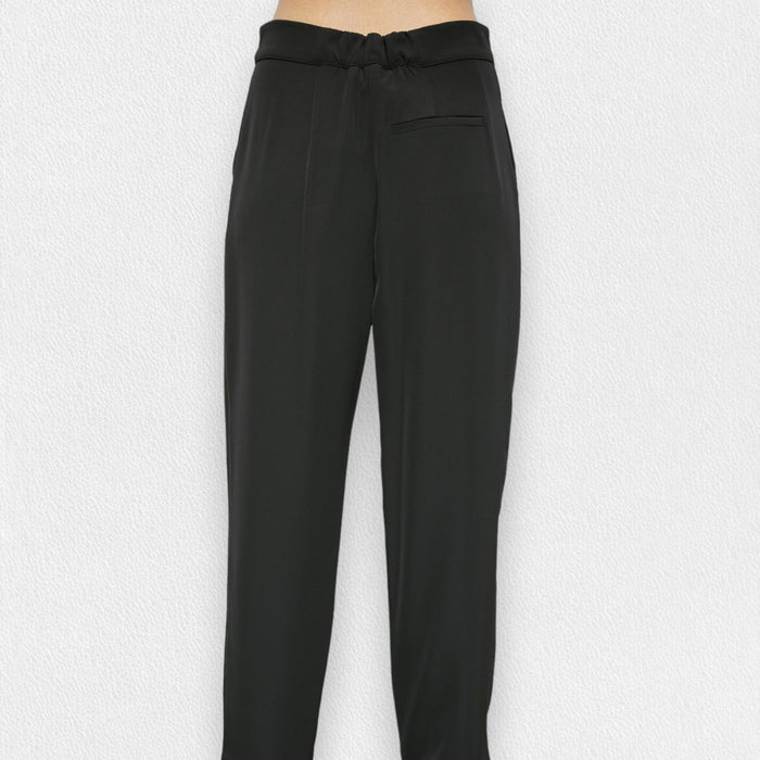 A person wears the MEIMEIJ DRAWSTRING TROUSER PANT—high-waisted, loose-fitting black pants in soft polyester with a tie belt—standing barefoot against a light textured background.