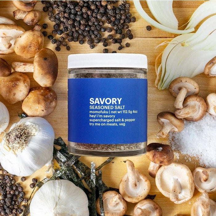 A clear jar with a white lid holds brown MOMOFUKU - SAVORY SALT, featuring a blue label with an orange fruit graphic and "Momofuku Savory Seasoned Salt" text. Net weight: 3.9 oz (112g). Made with premium kosher salt. Brand: MOMOFUKU.