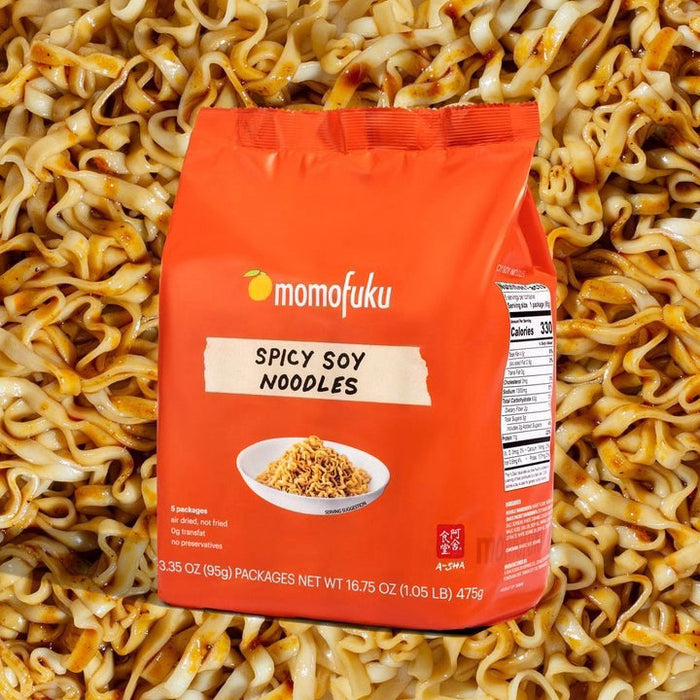A bright orange bag of MOMOFUKU - SPICY SOY NOODLES shows a bowl of air-dried noodles, notes five packs inside, no artificial ingredients, and a net weight of 16.75 oz (1.05 lb).
