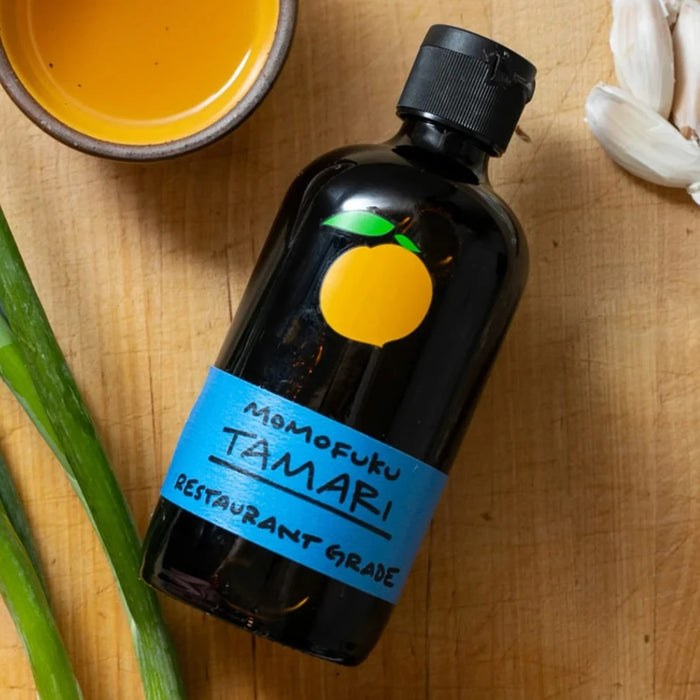 A dark glass bottle with a black cap and blue label reading "MOMOFUKU - TAMARI"—a premium, gluten-free soy sauce by MOMOFUKU—features an orange fruit graphic above the text. The bottle stands on a white surface.