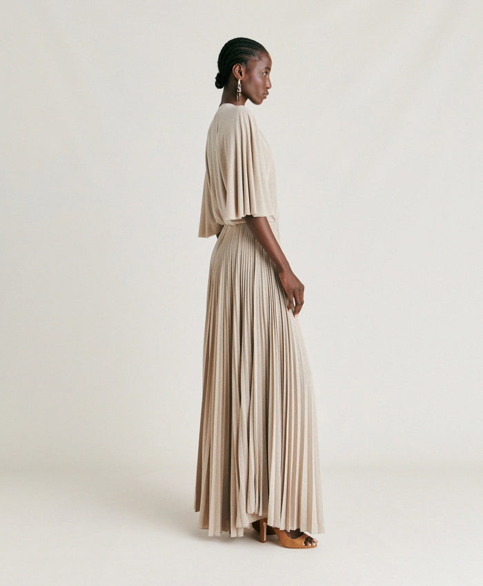 A woman poses against a plain background wearing the MOMONI - PARADISU DRESS by MOMONI, featuring light beige lurex jersey, kimono sleeves, and a pleated skirt. She styles it with braided hair, earrings, and open-toed shoes.
