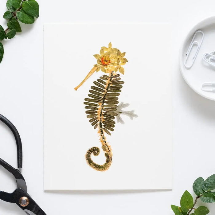 The PETAL PEOPLE PRESS - SEAHORSE CARD features a seahorse design made from pressed flowers and leaves, with green leaves for the body and yellow-brown flowers for the head and tail, set against a light wooden background.