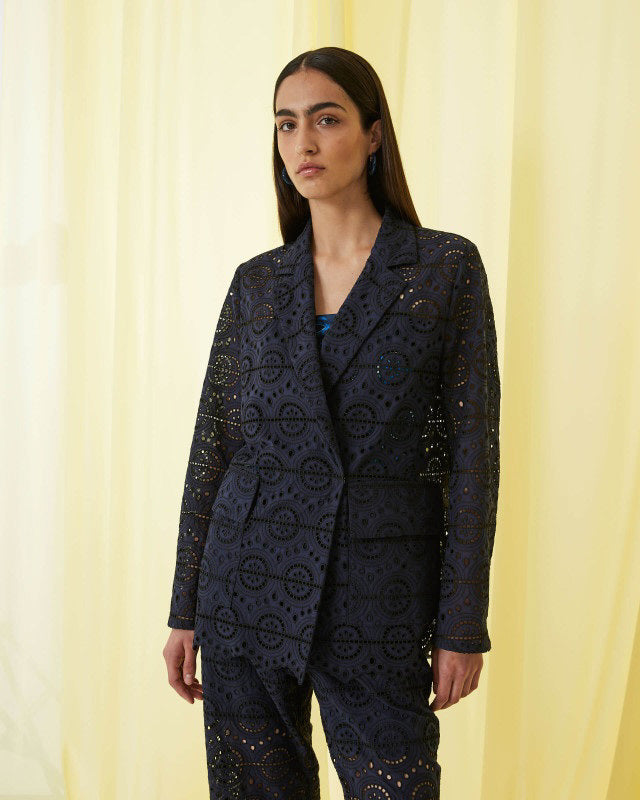 A woman with long dark hair wears the SFIZIO Double Breasted Broderi Anglaise Jacket by SFIZIO, a navy blue lace pantsuit with a geometric eyelet pattern, standing before yellow curtains.