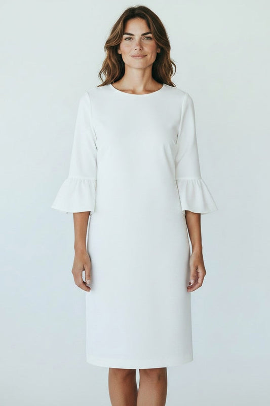 A woman with wavy brown hair smiles softly, facing forward in a knee-length 0039 ITALY - MAXLIN DRESS featuring three-quarter trumpet sleeves and ruffled cuffs. The white dress by 0039 ITALY looks comfortable against a plain light background.