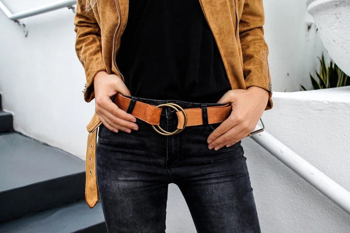 A person wearing a tan suede jacket, black shirt, and dark jeans stands on stairs, showcasing a tan belt with a large round gold buckle. Their hands rest on their hips.