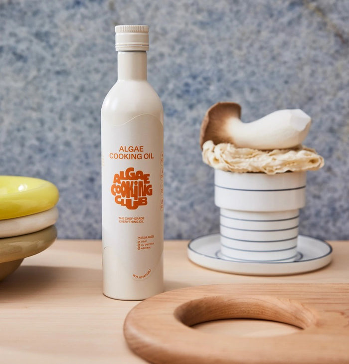 A white 16 fl oz bottle of ALGAE COOKING CLUB - ALGAE COOKING OIL is displayed, featuring orange text and graphics, including the stylized “Algae Cooking Club” branding. Designed for high-heat cooking and heart health, the bottle has a white cap and is set against a plain white background.