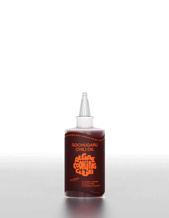 A clear plastic bottle labeled "ALGAE COOKING CLUB - GOCHUGARU CHILI OIL" in bold orange text sits on a white surface against a plain background, holding vibrant Korean chili oil from ALGAE COOKING CLUB.