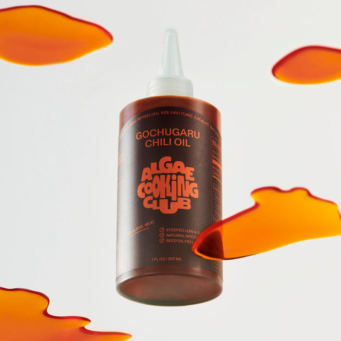 A clear plastic bottle labeled "ALGAE COOKING CLUB - GOCHUGARU CHILI OIL" in bold orange text sits on a white surface against a plain background, holding vibrant Korean chili oil from ALGAE COOKING CLUB.