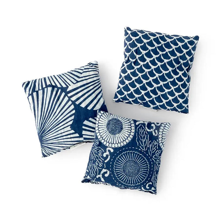 A person rests on a bed with white pillows, while in the foreground, a black and white cup sits on a saucer with lemon, mint, and Egyptian Chamomile. Nearby are BAUDELAIRE - CHAMOMILE SLEEP SACHETS and folded blue patterned cloths, enhancing the serene setting.
