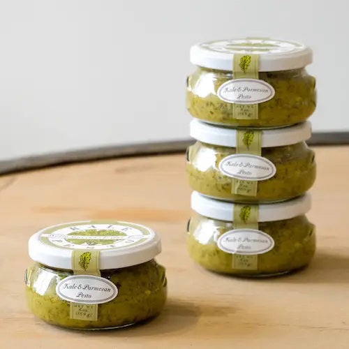 A small, round jar with a white lid labeled "BELLA CUCINA - KALE & PARMESAN PESTO." The jar is filled with green, all-natural pesto sauce and features a decorative label displaying the product name and net weight of 6 ounces (169 grams).