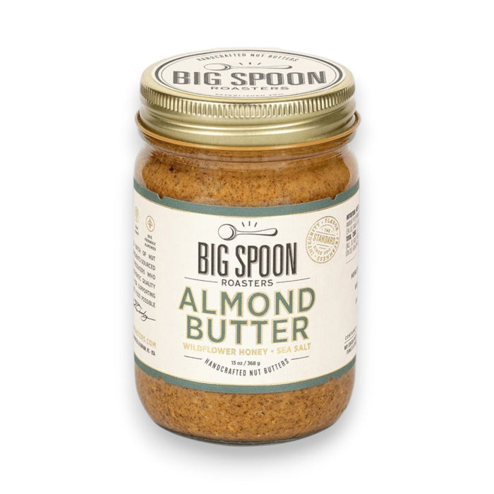 A glass jar of BIG SPOON ROASTERS - ALMOND BUTTER WILDFLOWER HONEY NUT BUTTER with a gold lid and a white label featuring branding and product details in black and gold text.