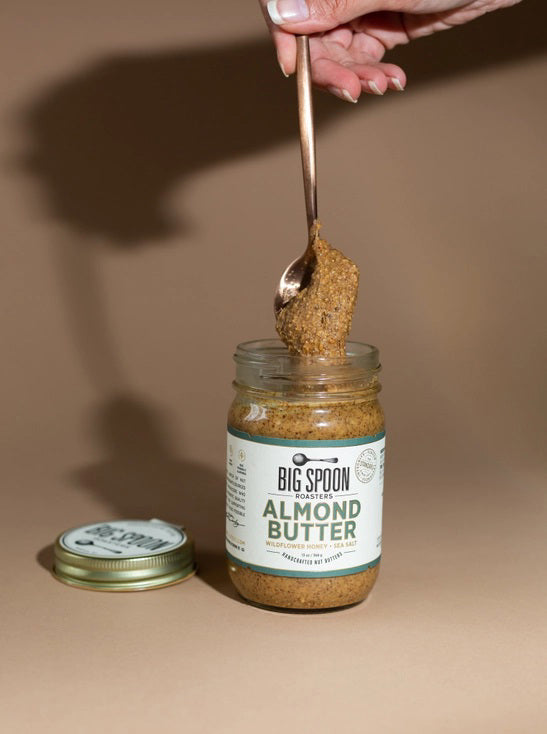 A glass jar of BIG SPOON ROASTERS - ALMOND BUTTER WILDFLOWER HONEY NUT BUTTER with a gold lid and a white label featuring branding and product details in black and gold text.