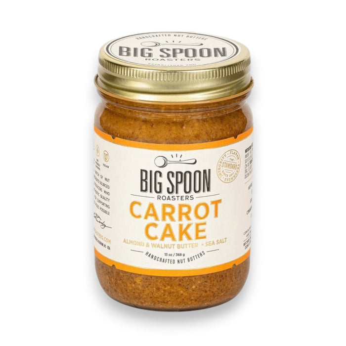 A jar of BIG SPOON ROASTERS - CARROT CAKE 13OZ, a vegan almond and walnut butter with no refined sugar, sits with a spoon inside in front of a frosted carrot cake and a plate holding a slice of the cake.