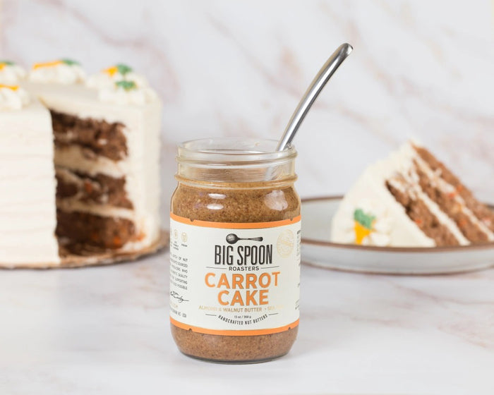 A jar of BIG SPOON ROASTERS - CARROT CAKE 13OZ, a vegan almond and walnut butter with no refined sugar, sits with a spoon inside in front of a frosted carrot cake and a plate holding a slice of the cake.