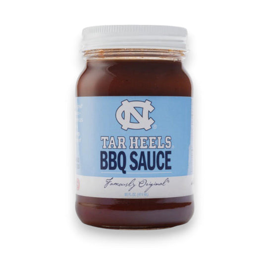 A jar of Cackalacky - Carolina Tar Heels BBQ Sauce is placed on a wooden surface, alongside a light blue football helmet and football. The display is artistically enhanced with scattered blue and white paper shreds, highlighting the vibrant appeal of this savory tomato-based barbecue sauce from Cackalacky.