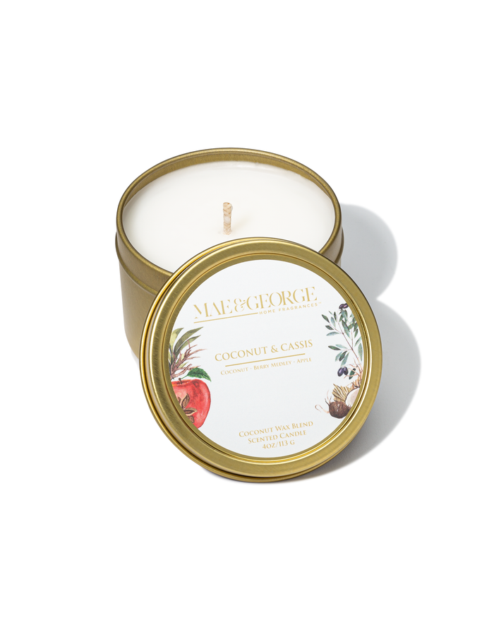 A gold travel candle with a label reading 'Mae & George Coconut & Cassis', along with images of coconuts and cassis berries. The candle is placed on a white surface.