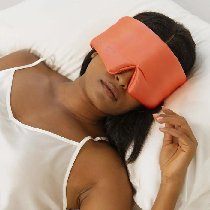 An older person with long gray hair wears a luxurious beige DROWSY - SATIN SLEEP MASK by DROWSY SLEEP CO, made of mulberry silk. In a matching satin top, they stand against a plain white background, ready for beauty sleep.