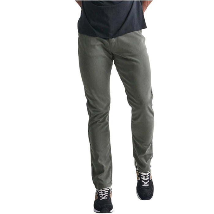 A person stands against a plain white background wearing DUER No Sweat Relaxed Taper Pants in Gull (olive green), a black T-shirt, and black New Balance sneakers with white soles and gold accents.