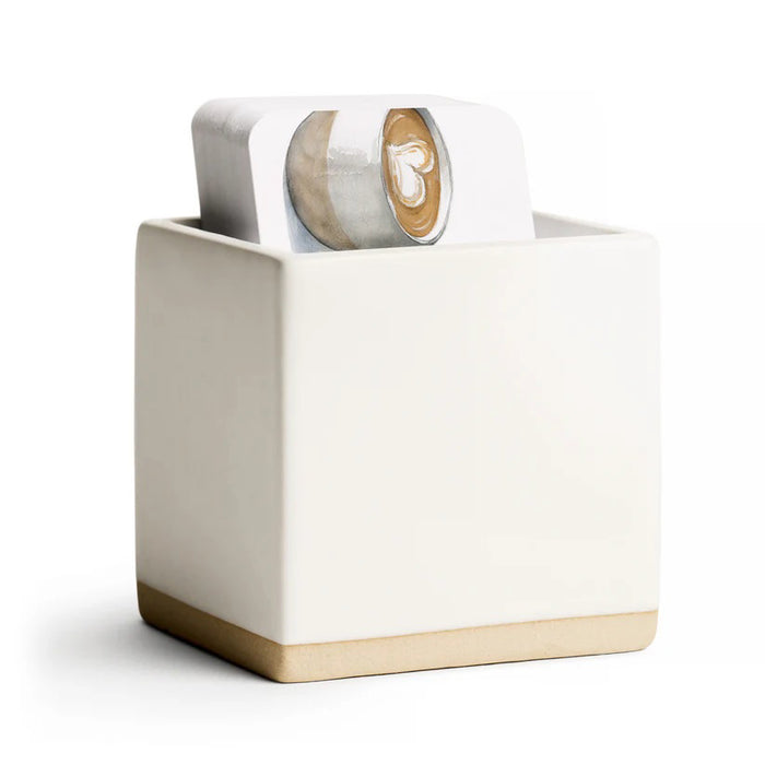 The E. FRANCES PAPER - LITTLE NOTES CERAMIC HOLDER - WHITE, designed as a modern minimalist ceramic cube gift box, contains decorative shredded paper and a gingerbread man-shaped white object, with the partially opened container revealing more shredded paper spilling out.