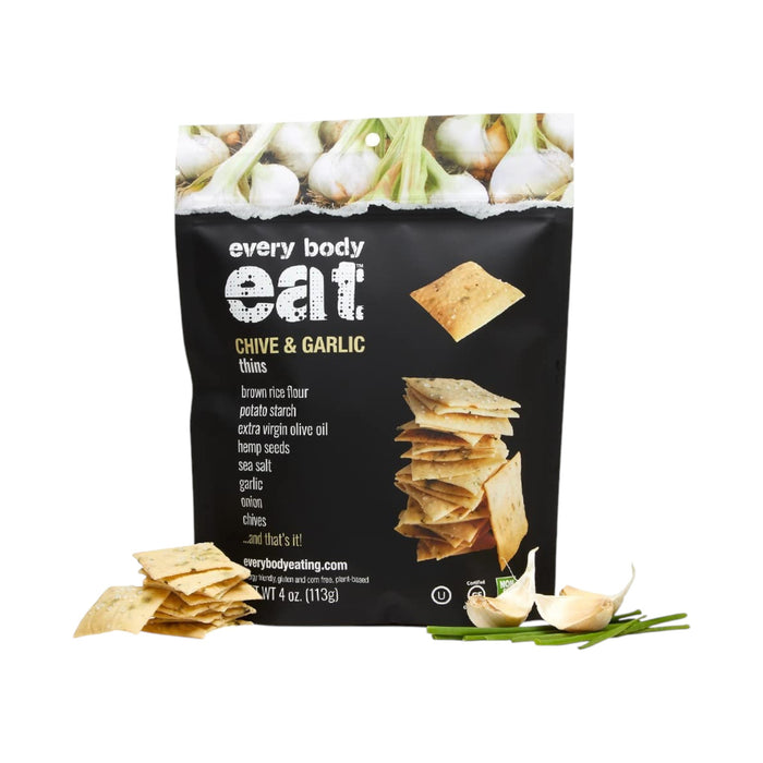A bag of EVERY BODY EAT CHIVE & GARLIC SNACK THINS from EVERY BODY EAT is pictured alongside gluten-free crackers, garlic cloves, and chives. The black packaging features images of crackers and garlic bulbs at the top, a list of ingredients, and highlights these allergy-friendly snacks.