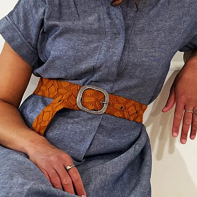 The JENNY KRAUSS - TERRACOTTA EMBROIDERED WOOL BELT, handcrafted in Peru, features a diamond embroidery pattern, metal buckle, and textured brown wool fabric, neatly coiled on a white surface.