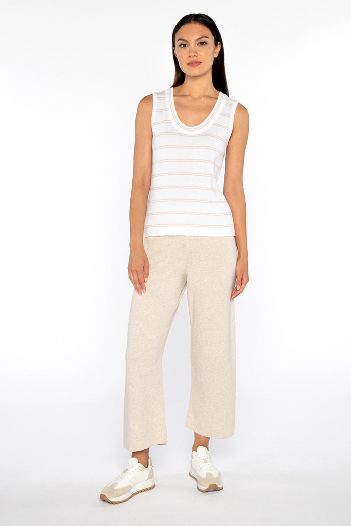 A woman with straight dark hair and a neutral expression stands against a white background, wearing a sleeveless white striped top, white sneakers, and the KINROSS CASHMERE Coverstitch Crop Cotton Blend Pant in light beige from KINROSS.