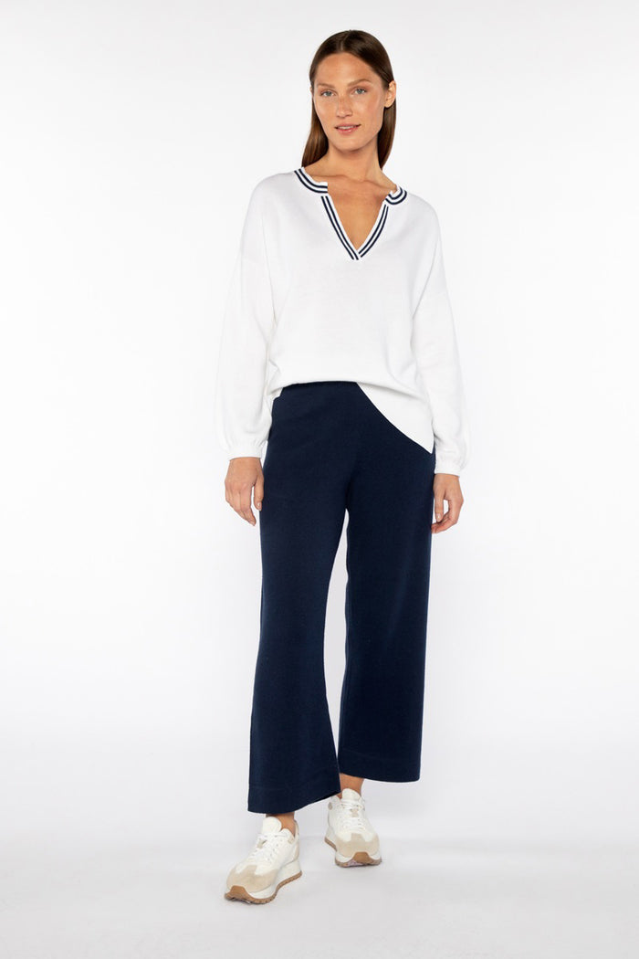 A woman with straight dark hair and a neutral expression stands against a white background, wearing a sleeveless white striped top, white sneakers, and the KINROSS CASHMERE Coverstitch Crop Cotton Blend Pant in light beige from KINROSS.