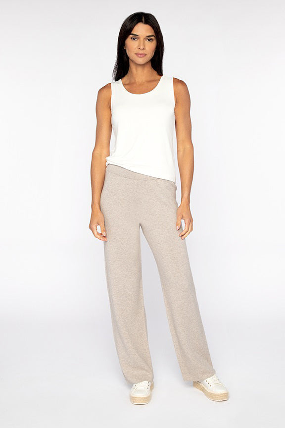 A woman stands against a plain white background, wearing loose light beige pants—the KINROSS CASHMERE Doubleknit Cotton Cashmere Pant by KINROSS—paired with a sleeveless white top and white sneakers. With long dark hair and a relaxed expression, her natural stance highlights these softly tailored pants as an understated addition to any elegant wardrobe.