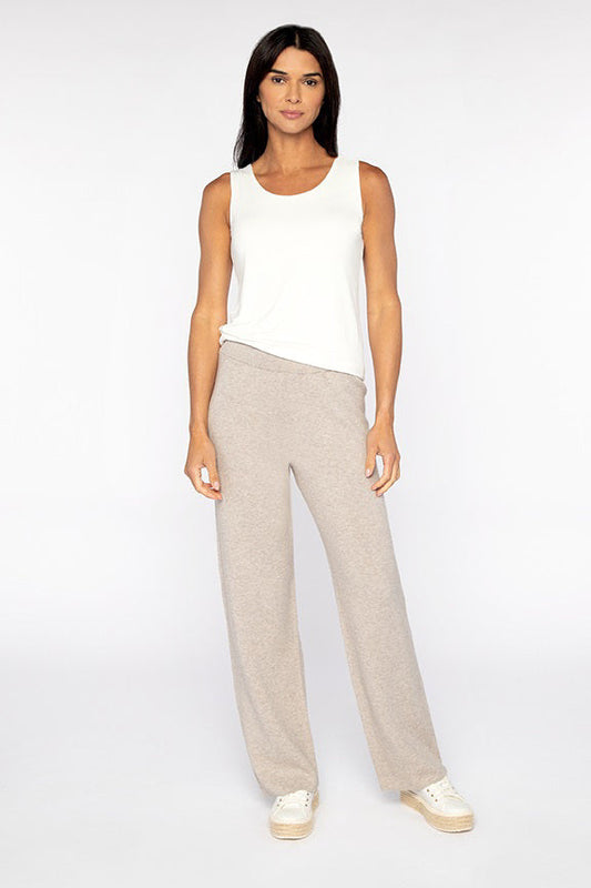A woman stands against a plain white background, wearing loose light beige pants—the KINROSS CASHMERE Doubleknit Cotton Cashmere Pant by KINROSS—paired with a sleeveless white top and white sneakers. With long dark hair and a relaxed expression, her natural stance highlights these softly tailored pants as an understated addition to any elegant wardrobe.