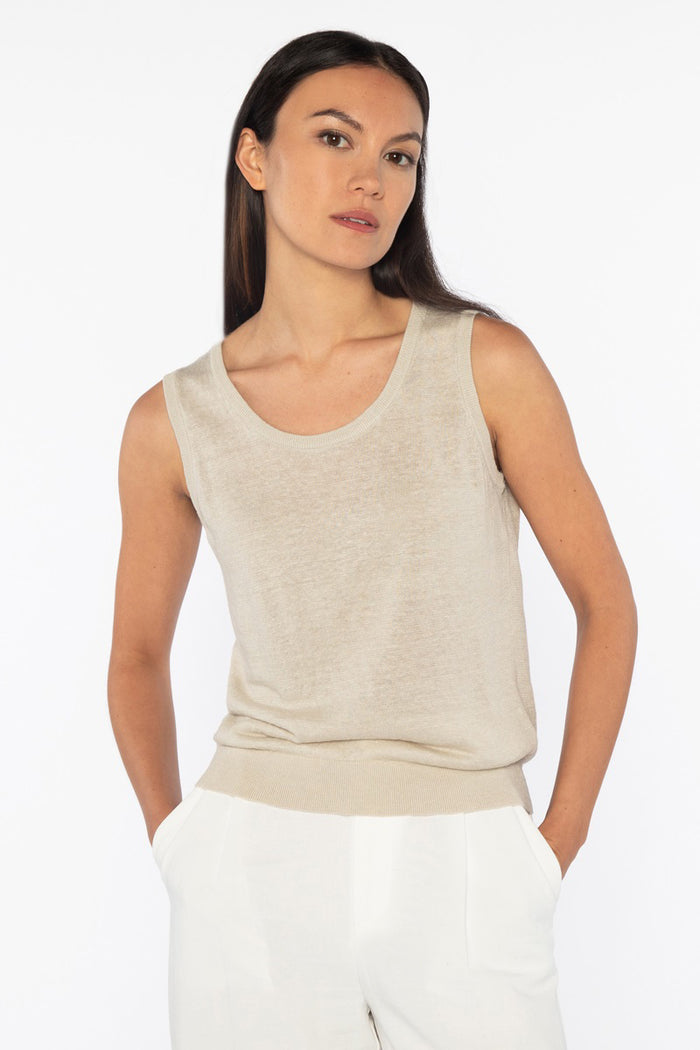 A woman with long dark hair models the KINROSS SLEEVELESS TANK IN LINEN, styled with white pants and hands in her pockets against a plain white background for a relaxed, everyday look.