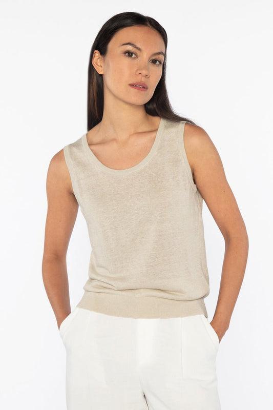 A woman with long dark hair models the KINROSS SLEEVELESS TANK IN LINEN, styled with white pants and hands in her pockets against a plain white background for a relaxed, everyday look.