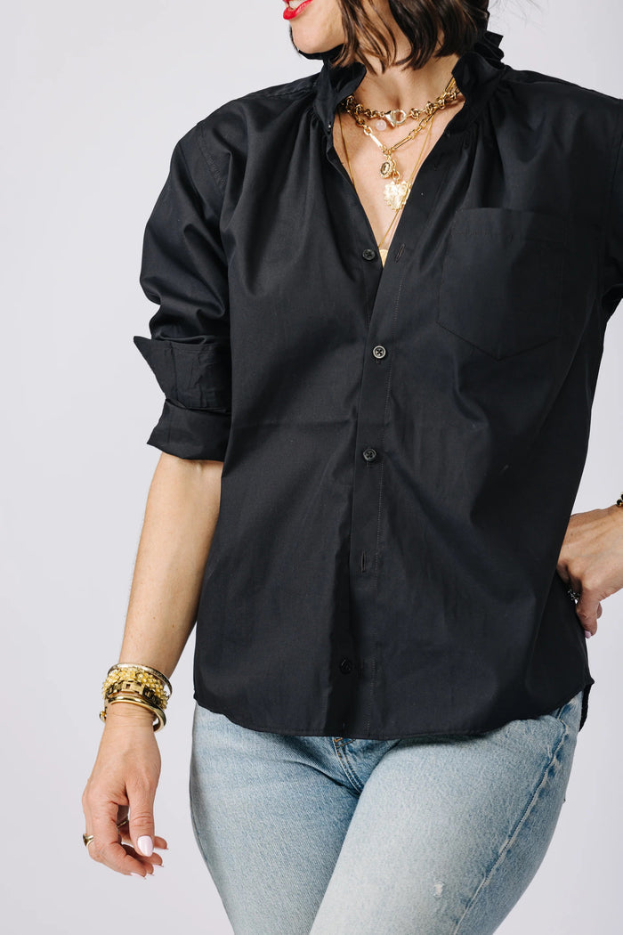 A woman with wavy dark hair and red lipstick leans against a wall outdoors, smiling at the camera. She wears the KMJ - QUARANTINA SHIRT by KMJ, sequined green pants, and gold jewelry. Green foliage and a sidewalk are behind her.