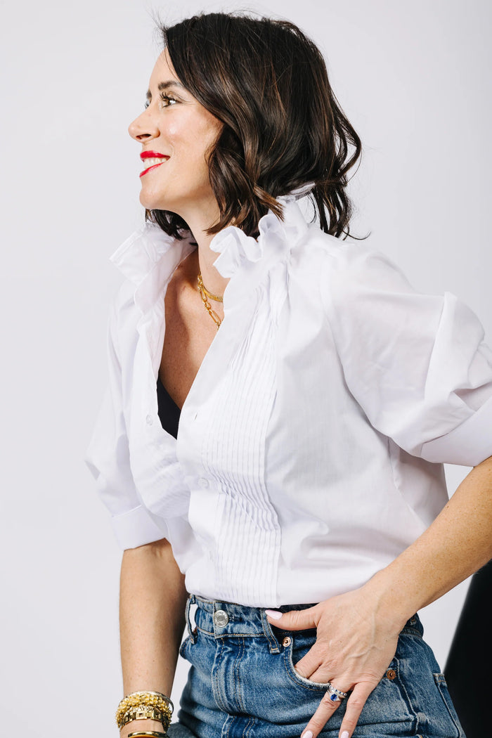 A blonde older woman wears the KMJ - SOUTHERN LOVER TUX blouse by KMJ and blue demi-boyfriend jeans with a turquoise necklace, looking down and tucking her hand in her waistband, set against a plain white background.