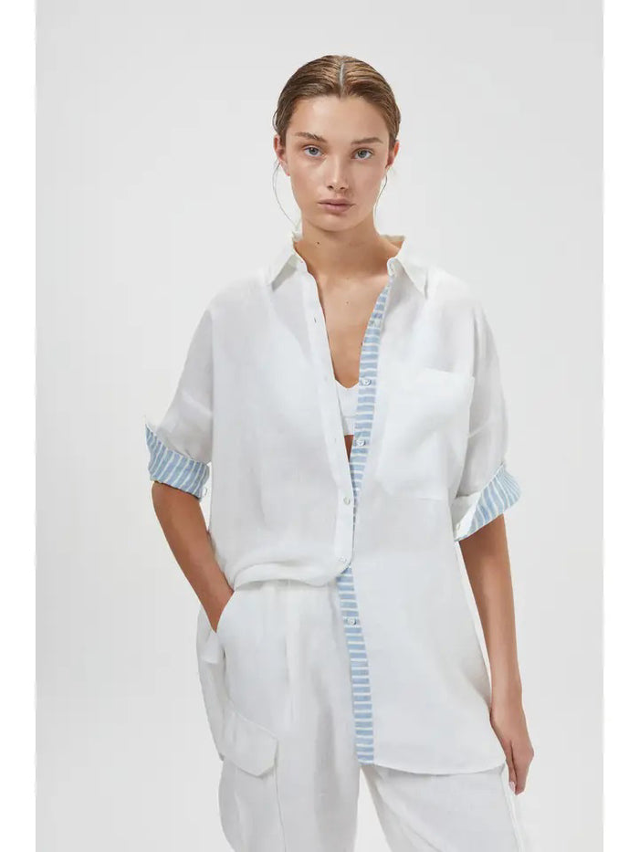 A woman wears the LANHTROPY ISLAND LINEN OVERSIZED SHIRT in white, featuring blue and white striped trim, paired with white pants. Her hair is tied back as she stands with one hand in her pocket against a plain light background.