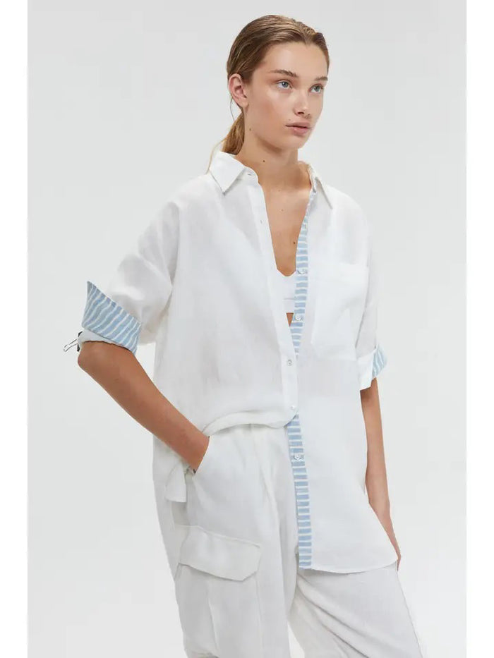 A woman wears the LANHTROPY ISLAND LINEN OVERSIZED SHIRT in white, featuring blue and white striped trim, paired with white pants. Her hair is tied back as she stands with one hand in her pocket against a plain light background.