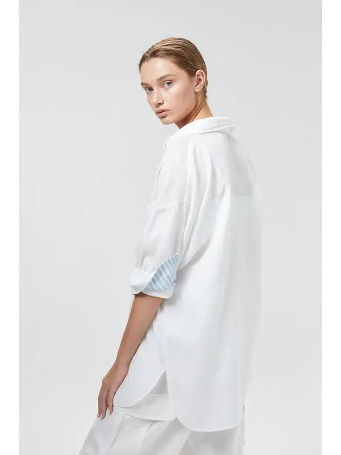 A woman wears the LANHTROPY ISLAND LINEN OVERSIZED SHIRT in white, featuring blue and white striped trim, paired with white pants. Her hair is tied back as she stands with one hand in her pocket against a plain light background.