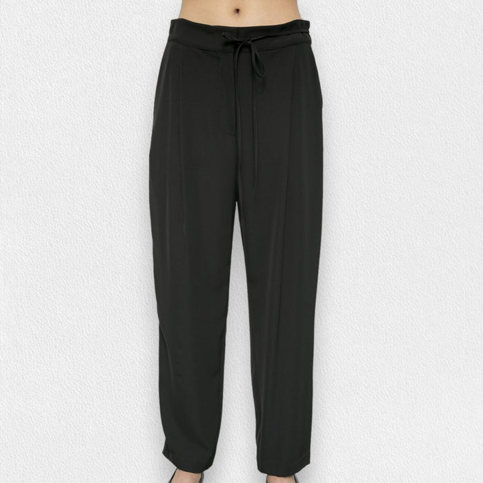 A person wears the MEIMEIJ DRAWSTRING TROUSER PANT—high-waisted, loose-fitting black pants in soft polyester with a tie belt—standing barefoot against a light textured background.