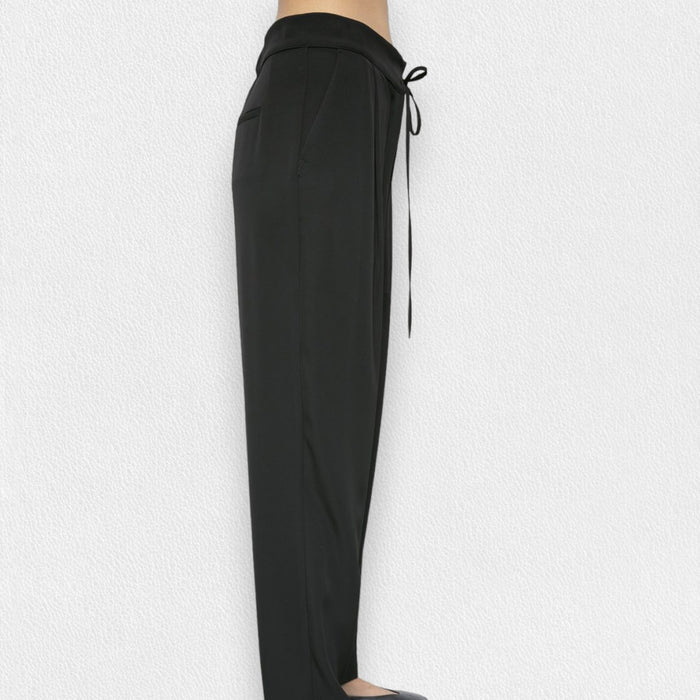 A person wears the MEIMEIJ DRAWSTRING TROUSER PANT—high-waisted, loose-fitting black pants in soft polyester with a tie belt—standing barefoot against a light textured background.