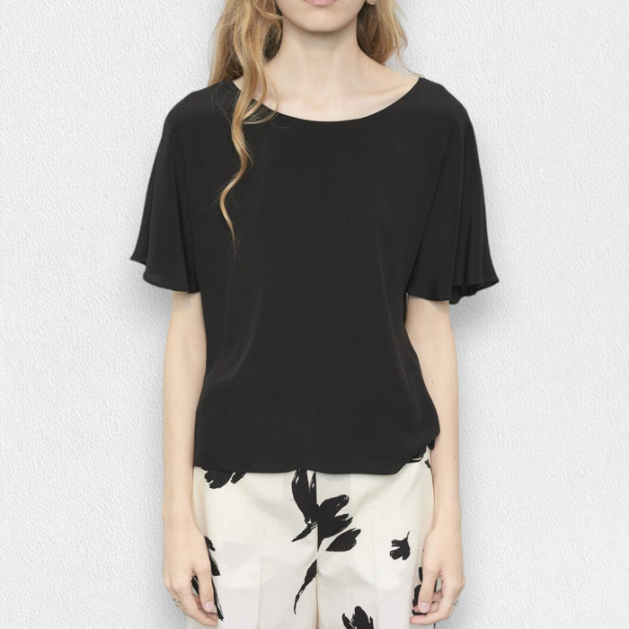 A woman with long, wavy blonde hair models the MEIMEIJ FLUTTER SLEEVE BLOUSE, paired with bold black floral print pants. The refined look is effortless and perfect for washable silk styles, set against a light textured background.