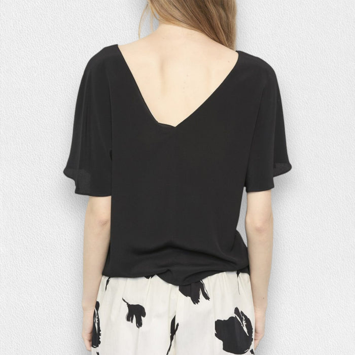 A woman with long, wavy blonde hair models the MEIMEIJ FLUTTER SLEEVE BLOUSE, paired with bold black floral print pants. The refined look is effortless and perfect for washable silk styles, set against a light textured background.