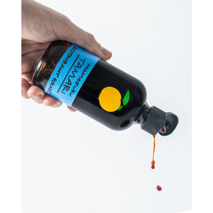 A dark glass bottle with a black cap and blue label reading "MOMOFUKU - TAMARI"—a premium, gluten-free soy sauce by MOMOFUKU—features an orange fruit graphic above the text. The bottle stands on a white surface.