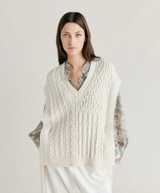 A woman with straight brown hair wears the MOMONI - ARENE KNIT VEST by MOMONI, a cream cable knit sweater vest made in Italy, styled over a patterned shirt and white pants, standing against a neutral background.