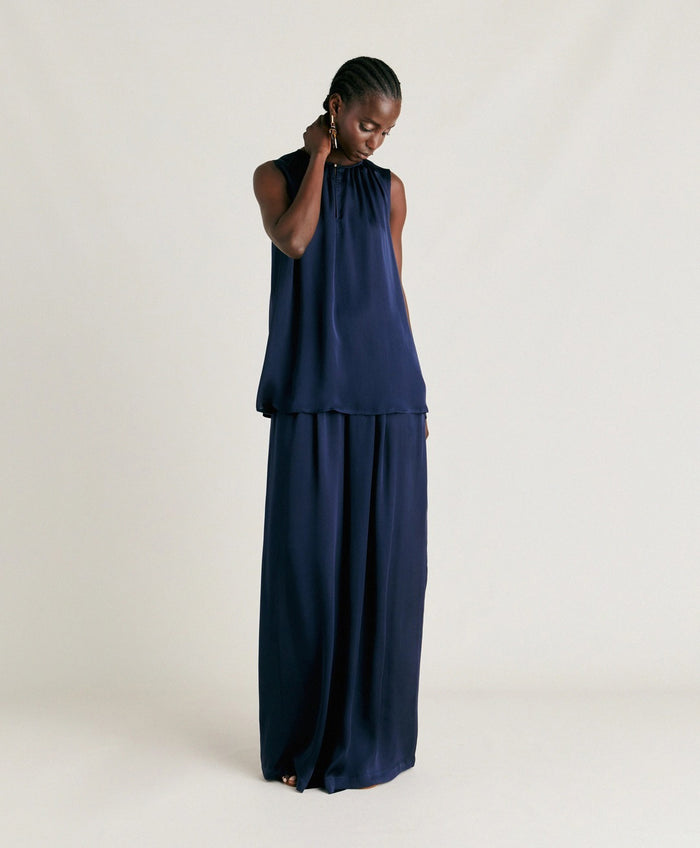 A person stands against a plain light background, wearing the MOMONI - CRIS SLEEVELESS TOP in dark navy silk with a round neck and a shiny finish. They are looking down and gently touching their ear. Brand: MOMONI.