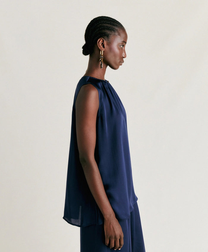 A person stands against a plain light background, wearing the MOMONI - CRIS SLEEVELESS TOP in dark navy silk with a round neck and a shiny finish. They are looking down and gently touching their ear. Brand: MOMONI.