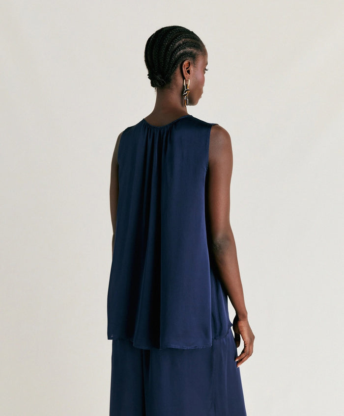 A person stands against a plain light background, wearing the MOMONI - CRIS SLEEVELESS TOP in dark navy silk with a round neck and a shiny finish. They are looking down and gently touching their ear. Brand: MOMONI.
