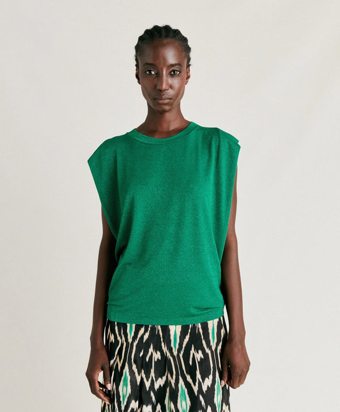 A woman with braided hair wears the MOMONI - ENNA LUREX JERSEY TOP, a sleeveless green top by MOMONI, paired with a black, white, and green 80s-inspired patterned skirt against a plain light background.
