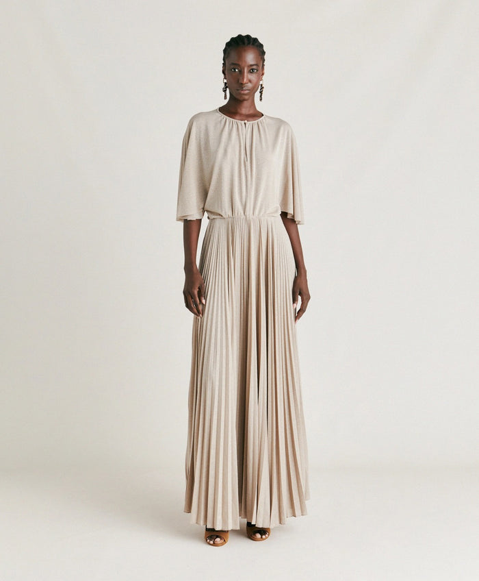 A woman poses against a plain background wearing the MOMONI - PARADISU DRESS by MOMONI, featuring light beige lurex jersey, kimono sleeves, and a pleated skirt. She styles it with braided hair, earrings, and open-toed shoes.