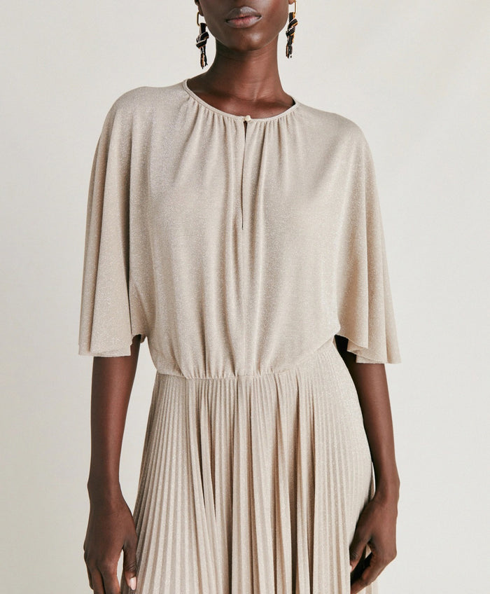 A woman poses against a plain background wearing the MOMONI - PARADISU DRESS by MOMONI, featuring light beige lurex jersey, kimono sleeves, and a pleated skirt. She styles it with braided hair, earrings, and open-toed shoes.