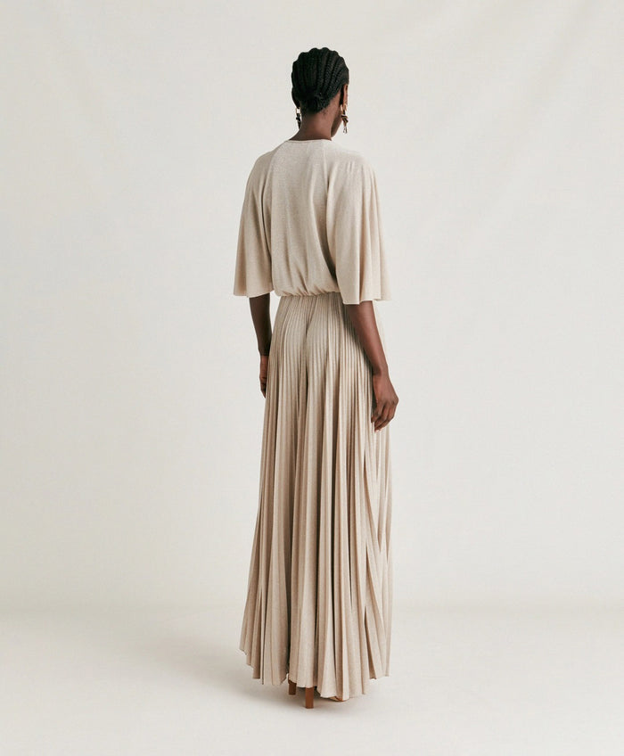 A woman poses against a plain background wearing the MOMONI - PARADISU DRESS by MOMONI, featuring light beige lurex jersey, kimono sleeves, and a pleated skirt. She styles it with braided hair, earrings, and open-toed shoes.